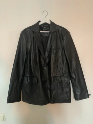 Svart skinnjacka från Bershka - Stilren svart skinnjacka från Bershka med klassisk krage och två knappar framtill. Jackan har raka ärmar, fickor på sidorna och en bröstficka. Perfekt för dig som vill ha en tidlös och trendig look.