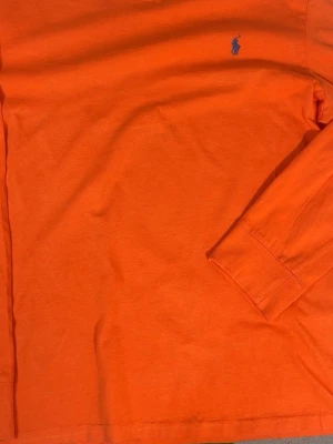 Orange långärmad tröja Polo Ralph Lauren - Snygg orange långärmad tröja från Polo Ralph Lauren med klassisk blå logga broderad på bröstet. Tröjan har rund halsringning och är gjord i mjuk bomull som känns skön mot huden. Perfekt för dig som vill ha en enkel men stilren look.