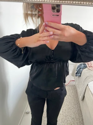 Svart satinblus med v-ringning -  svart blus i glansigt satinmaterial med v-ringning och knappar framtill, resår i midjan och vida, långa ärmar med puff🤗säljer även bikbok vera kostymbyxorna på bilden💕