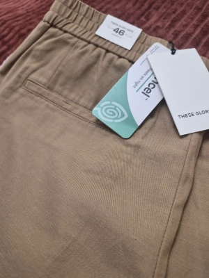 These GLORY DAYS T Amalfi Tencel beige byxor, storlek 46 - Snygga beige mjukisbyxor från These Glory Days med elastisk midja och raka ben. Byxorna har diskreta sidofickor och är tillverkade i ett mjukt, vävt material som känns skönt mot huden. Perfekta för en avslappnad och stilren look.