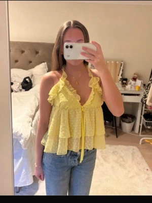 Gul volang topp med knyt i nacken - Söt gul topp med volanger och små prickar, knyt i nacken och öppen rygg. Luftig och perfekt för sommaren. Toppen har v-ringning och är ärmlös med ett lätt transparent material.