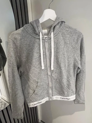 Grå hoodie med dragkedja Calvin Klein. S - Grå hoodie från Calvin Klein med dragkedja, vita snören i huvan och två fickor fram. Mjuk bomullsblandning och vit kant med Calvin Klein-logga längst ner. Den är i storlek S💖