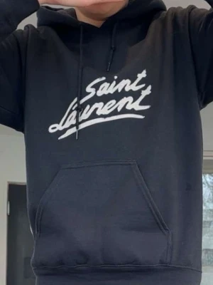 Svart Saint Laurent hoodie - Snygg svart hoodie från Saint Laurent med vit logga framtill i handskriven stil. Klassisk känguruficka och justerbar huva med snören. Perfekt för dig som gillar streetwear och vill ha en stilren look.