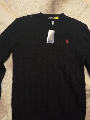 Svart kabelstickad tröja Polo Ralph Lauren - Kabelstickad svart tröja från Polo Ralph Lauren med rund halsringning och röd broderad logga på bröstet. Tröjan har långa ärmar och ribbade muddar vid ärmslut och nederkant. Perfekt för dig som gillar klassisk och stilren design.