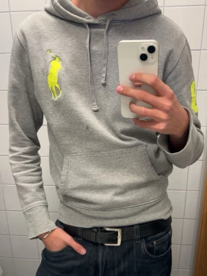 Ralph Lauren hoodie - Snygg grå hoodie från Polo Ralph Lauren med stor gul logga på bröstet och siffran 3 i gult på ärmen. Tillverkad i en mjuk bomullsblandning med huva och känguruficka framtill. Perfekt för en avslappnad och trendig stil.