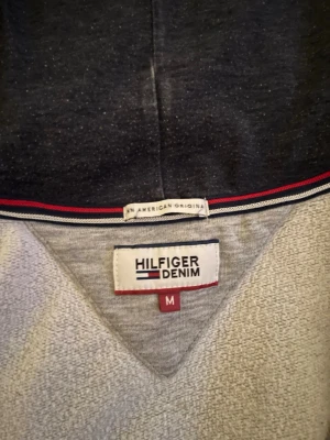 Grå Hilfiger Denim hoodie med dragkedja - Snygg grå hoodie från Hilfiger Denim i storlek M. Hoodien har dragkedja framtill, två fickor och en klassisk huva med mörkblå insida. Detaljer med Hilfigers logga på bröstet och röd/vit/blå kantband vid huvan. Perfekt för en avslappnad stil.