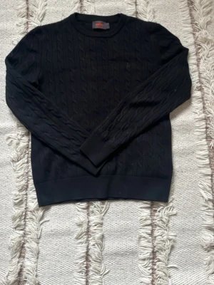Svart kabelstickad tröja - Kabelstickad svart tröja med rund halsringning och långa ärmar. Tröjan har ribbade muddar vid ärmslut och nederkant, vilket ger en klassisk och stilren look. Perfekt att matcha med jeans eller kjol för en enkel men snygg outfit.