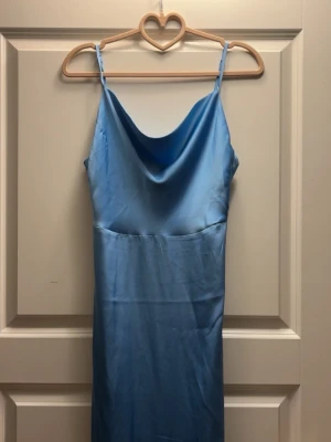 Balklänning  - Säljer en elegant ljusblå balklänning med tunna axelband och draperad urringning. Klänningen har en slät, glansig yta och är figurnära i modellen. Perfekt för dig som vill ha en stilren och trendig look. 