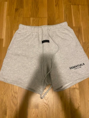 Grå Essentials Fear of God shorts - Säljer ett par ljusgrå shorts från Fear of God Essentials med snörning i midjan och tryckt logga på benet. Sköna och stilrena, perfekta för sommaren eller chill hemma. Klassisk passform och mjukt material.