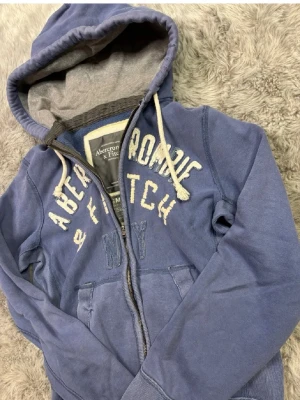 Blå hoodie från Abercrombie & Fitch - Säljer en blå hoodie från Abercrombie & Fitch med dragkedja och vit broderad logga på bröstet. Hoodien har grått innerfoder i huvan och vita snören. Perfekt för en avslappnad och cool stil. (Storlek S)