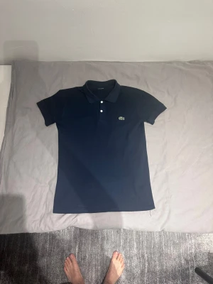 Mörkblå piké från Lacoste - Snygg mörkblå pikétröja från Lacoste med klassisk krage och två knappar framtill. Ikoniska krokodilloggan på bröstet. Tillverkad i mjuk bomull och har korta ärmar för en clean och stilren look.