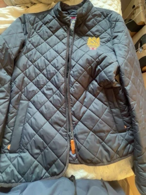 Svart quiltad vindjacka Morris - Snygg svart quiltad vindjacka från Morris med gul broderad logga på bröstet. Jackan har ståkrage, dragkedja framtill och två fickor. Tillverkad i ett lätt och slitstarkt material som passar perfekt till kyliga dagar.