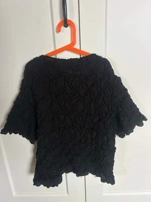 Svart crinkle topp med kort ärm - Säljer en svart topp med crinkle-effekt och korta ärmar. Toppen har en lös passform och vågig kant nertill och vid ärmsluten. Perfekt för dig som gillar en unik struktur och enkel stil. Kan vara lite kort i längden.