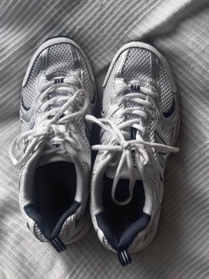 New Balance vita sneakers - Säljer ett par vita New Balance sneakers med mesh och syntetmaterial. Skorna har klassisk låg profil, vita snören och detaljer i mörkblått. Perfekta för dig som gillar sportig och clean stil.