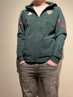 Adidas Manchester United träningsjacka - Mörkgrön träningsjacka från Adidas med Manchester United-logga på bröstet. Jackan har hel dragkedja, huva, vita Adidas-detaljer och röda ränder längs ärmarna. Tillverkad i ett sportigt material med ribbade muddar för extra komfort.