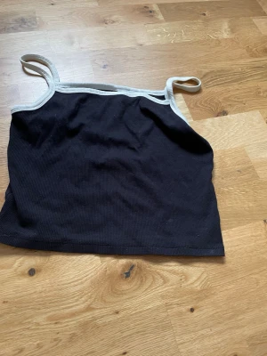 Svart ribbad linnetopp från H&M - Snygg svart ribbad linnetopp från H&M med vita kontrastkanter och smala axelband. Perfekt för en enkel och stilren look. Tillverkad i mjuk bomull som känns skön mot huden.