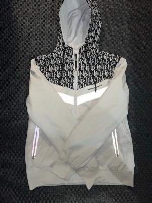 Vindjacka  - Snygg vit vindjacka från Black Squad med svartvit mönstrad huva, Jackan har dragkedja framtill, två fickor med reflekterande detaljer och elastiska muddar vid ärmslut och nederkant. Vindjackan har även reflekterande detaljer vid ovansidan av jackan, detta är ett måste! Perfekt för dig som vill ha en stilren och modern look.