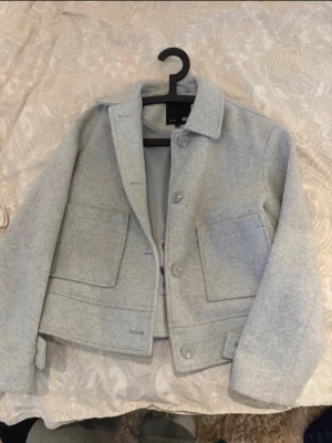 Ljusgrå ulljacka från Lager 157 - Stilren ljusgrå jacka från Lager 157 i ullmix med rak passform. Jackan har stora framfickor, klassisk krage och stängs med knappar framtill. Perfekt för lager på lager och enkel att matcha med olika outfits.