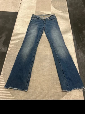 Blå bootcut jeans från Nelly - Säljer ett par blå bootcut jeans från Nelly med råa, fransiga kanter i midjan och vid bensluten. Jeansen har en klassisk femficksmodell och slitningar framtill för en cool vintagekänsla. Perfekta för dig som gillar en avslappnad och trendig stil.