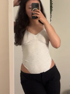 Ljusgrå body med tunna axelband - Säljer en enkel och stilren ljusgrå body med tunna axelband och lätt rynkning vid bysten. Bodyn har en mjuk, stretchig passform och är perfekt att bära under andra plagg. Materialet känns bekvämt mot huden och ger en smickrande siluett.