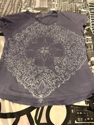 Lila t-shirt med mandalamönster - Säljer en lila t-shirt med korta ärmar och ett stort vitt mandalainspirerat tryck på framsidan. T-shirten har rund halsringning och är gjord i mjukt bomullsmaterial. Perfekt för dig som gillar unika och snygga prints! Finns ingen storlek på den. Men för referens, har jag XL/L i storlek för tröjor och den är lite tajt på mig, den kan vara L/M någonstans där. 