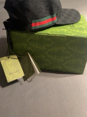 Svart Gucci keps med GG-mönster - Snygg svart keps från Gucci med det klassiska GG-monogrammet över hela kepsen. På sidan finns en grön och röd rand som ger en ikonisk touch. Kepsen har böjd skärm och justerbar rem bak. Materialet är slitstark canvas som känns lyxig och trendig.