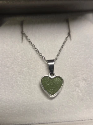 Halsband med grönt hjärthänge - Snyggt halsband med ett hjärtformat hänge i glittrande grönt som ändrar färg i mörker till ett blått sken. Effekten aktiveras av vanligt dagsljus utifrån och skenet varar vanligtvis i flera timmar. Kedjan är tunn och silverfärgad i rostfritt stål, 45 cm, vilket ger ett stilrent och modernt intryck. Perfekt accessoar för att lyfta din outfit med en touch av färg och personlighet. Tål vatten och missfärgas inte. Lång livslängd. 