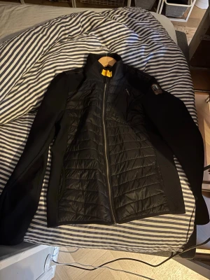 Svart jacka Parajumpers - Snygg svart jacka från Parajumpers med quiltade partier framtill och på ryggen. Jackan har hög krage, dragkedja hela vägen och en patch på axeln. Perfekt för kyliga dagar och har en sportig look.