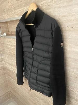 Moncler cardigan - Snygg svart pufferjacka från Moncler med quiltad front och rygg samt släta ärmar. Jackan har dragkedja framtill, två sidofickor med dragkedja och en liten Moncler-logga på ärmen. Perfekt för kalla vinterdagar och riktigt stilren look. Skriv vid frågor