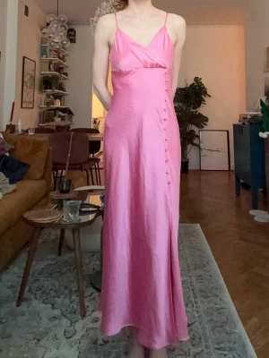 Rosa lång satinklänning med smala band - Säljer en elegant rosa midiklänning/långklänning i satin med smala axelband och omlott upptill. Klänningen har dekorativa knappar längs sidan och en figurnära passform som ger en snygg siluett. Perfekt för dig som gillar en stilren och trendig look. Klänningen har en slits. Banden är inte justerbara. Oanvänd. Tyvärr är materiallappen bortklippt. Perfekt nu till våren och sommaren till antingen fester, tillställningar, bröllop eller bara till vardags. Har ej stretch.