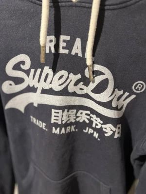Mörkblå Superdry hoodie med tryck - Snygg mörkblå hoodie från Superdry med stort vitt logotryck på bröstet och japanska tecken. Klassisk känguruficka framtill och vita dragsnören i huvan. Perfekt för en avslappnad streetwear-look.
