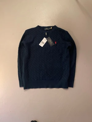 Mörkblå kabelstickad tröja Polo Ralph Lauren - Kabelstickad mörkblå tröja från Polo Ralph Lauren med rund halsringning och den klassiska röda logotypen broderad på bröstet. Tröjan har ribbade muddar och är tillverkad i mjukt material som ger en lyxig känsla.