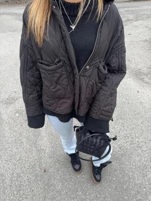 Svart quiltad vindjacka med huva - Snygg svart quiltad vindjacka med dragkedja och två stora fickor framtill. Jackan har en avslappnad passform, ribbade muddar vid ärmsluten och en praktisk huva. Perfekt för lager på lager och streetstyle.