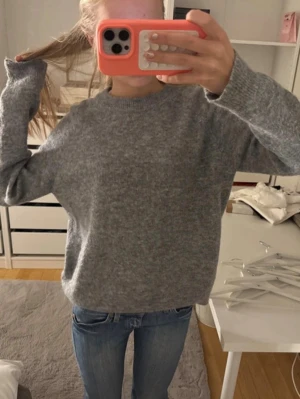 Grå stickad tröja i mjukt material - Mysig grå stickad tröja med rund hals och långa ärmar. Tröjan har en enkel, oversized passform och är gjord i ett mjukt material som känns skönt mot huden. Perfekt att bära till jeans för en avslappnad look.