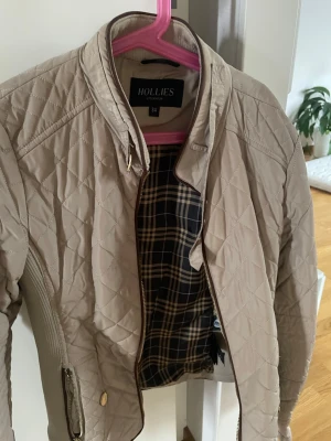 Beige quiltad jacka från Hollies - Snygg beige quiltad jacka från Hollies med rutigt innerfoder och dragkedja framtill. Jackan har hög krage och ribbade detaljer vid sidorna för extra komfort. Perfekt för dig som gillar klassisk men trendig stil. I storlek 34, knappt andvänd och i bra skick utan fläckar eller fel