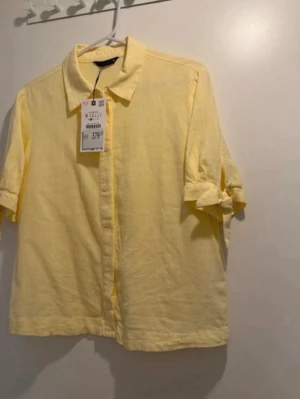 Gul kortärmad skjorta med knyt - Fräsch gul skjorta från Uniqlo med korta ärmar och snygg knyt-detalj på ärmarna. Skjortan har klassisk krage och knappar framtill, perfekt för dig som vill ha en enkel men trendig look. Materialet är linne som känns svalt och bekvämt.