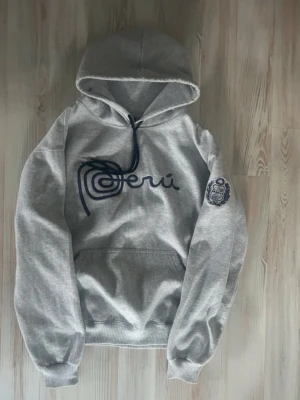 Grå hoodie med Peru-tryck - Snygg grå hoodie med stor huva och känguruficka. Framsidan har ett mörkblått Peru-tryck och ärmen är dekorerad med ett broderat emblem. Mjuk bomullsblandning och ribbade muddar för extra komfort. Perfekt för en avslappnad streetstyle. PRIS KAN DISKUTERAS 
