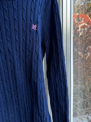 Mörkblå kabelstickad tröja Gina Tricot - Lik Ralph lauren! Mörkblå kabelstickad tröja från Gina Tricot med rund halsringning och ribbade muddar. Tröjan har en rosa broderad logga på bröstet och rosa detalj på insidan av kragen. 