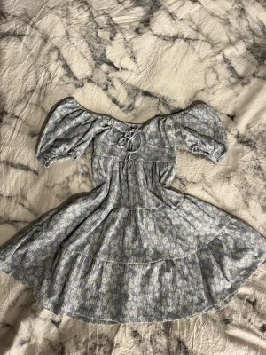 Blå blommig kortklänning med puffärm - Söt blå kortklänning med vitt blommönster och puffärmar. Klänningen har snörning framtill, omlottkänsla vid bysten och volangdetaljer i kjolen. Perfekt för sommarens alla tillfällen.