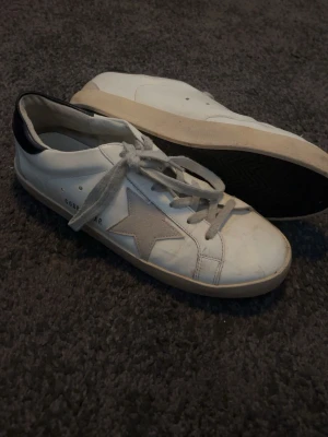 Golden Goose Superstars - Golden Goose sneakers i vitt läder med grå mockastjärna på sidan och svart hälparti. Klassisk låg modell med vita snören och beige sula. Märkeslogga på hälen och insidan. Perfekta för en avslappnad streetstyle-look.