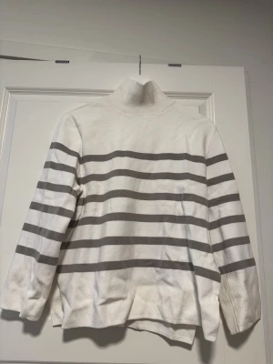 Vit stickad polotröja med grå ränder - Snygg vit stickad polotröja med breda horisontella grå ränder. Tröjan har långa ärmar, hög krage och slitsar i sidorna för en avslappnad look. Perfekt att matcha med jeans eller kjol för en trendig stil.