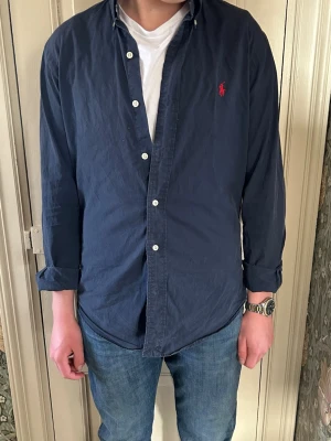 Mörkblå skjorta från Ralph Lauren - Klassisk mörkblå skjorta från Ralph Lauren med vit knappstängning och röd broderad logga på bröstet. Skjortan har lång ärm, button-down krage och är tillverkad i bomull. Perfekt till jeans för en clean och stilren look.