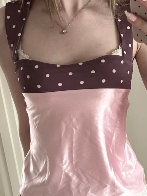 Rosa satin fest topp - Söt linnetopp i glansigt rosa material med bruna axelband och överdel dekorerad med ljusrosa prickar. Toppen har breda axelband och knytband i ryggen för en justerbar passform. Perfekt för dig som gillar unika detaljer och lekfull stil.