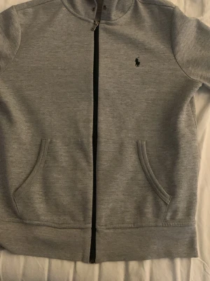 Grå zip hoodie från Polo Ralph Lauren - Snygg grå hoodie från Polo Ralph Lauren med dragkedja framtill och två fickor. Klassisk logga broderad på bröstet. Tillverkad i mjukt bomullsmaterial som känns skönt mot huden. Perfekt för en avslappnad och stilren look.         Självklart äkta STRLK M