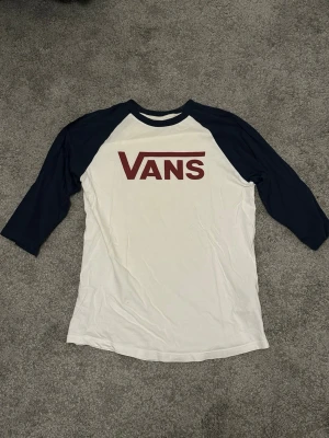 Vans baseballtröja med marinblå ärmar - Säljer en snygg baseballinspirerad t-shirt från Vans med marinblå trekvartsärmar och vit kropp. Stort rött Vans-tryck på bröstet. Perfekt för en avslappnad streetwear-look. Tillverkad i mjuk bomull för skön känsla.