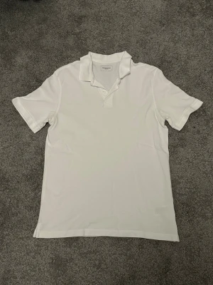 Vit pikétröja från Dressmann - Klassisk vit pikétröja från Dressmann med krage och två knappar framtill. Tröjan har korta ärmar och är tillverkad i ett mjukt bomullsmaterial som känns skönt mot huden. Enkel och stilren design som passar till det mesta.