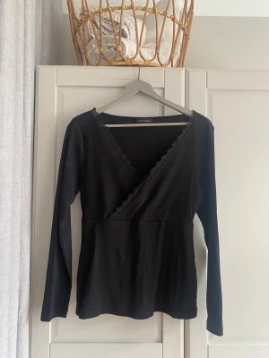 Svart omlottopp med spetsdetalj - Svart omlottopp från Anna Field med långa ärmar och v-ringning. Toppen har fina spetsdetaljer längs ringningen och är tillverkad i ett mjukt material som sitter snyggt. Perfekt att styla med jeans eller kjol.