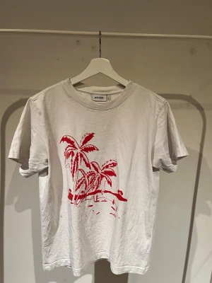 Vit t-shirt med palmtryck från Weekday - Säljer en vit t-shirt från Weekday med ett rött palmtryck framtill. Klassisk rund halsringning och korta ärmar. Tillverkad i mjuk bomull som är skön att bära. Perfekt för dig som gillar enkel men snygg stil.