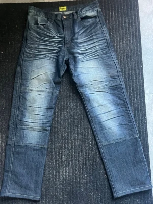 Blå motorcykel jeans med glidskydd  - Säljer ett par blå motorcykel jeans med snygga slitningar och tvättade detaljer framtill och baktill. Jeansen har raka ben och klassisk femficksmodell. Bakfickorna har broderad logga och materialet är kraftig denim. Perfekta för en avslappnad streetstil.