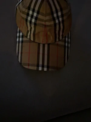 Burberry klassisk rutig keps - Säljer en ikonisk keps från Burberry med det klassiska beigea rutmönstret och svarta, vita samt röda detaljer. Kepsen har böjd skärm och justerbar passform. Materialet är bomull som känns både slitstarkt och bekvämt. Pris diskuteras